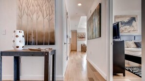 A modern interior with a wooden floor, a dining area with a table and chairs, a hallway leading to a bedroom with a bed and a TV. - Spacious 1BR | Full Kitchen | Washer/Dryer | Balcony | by GLS (New York)