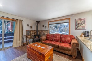 Uma sala de estar acolhedora com lareira de pedra, um sofá vermelho estampado, um baú de madeira e uma grande janela com vista para uma paisagem nevada.