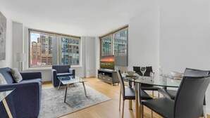 Una sala de estar moderna con sofá, una mesa de centro de vidrio, una mesa de comedor con sillas y televisor. Amplias ventanas ofrecen vistas de edificios exteriores.