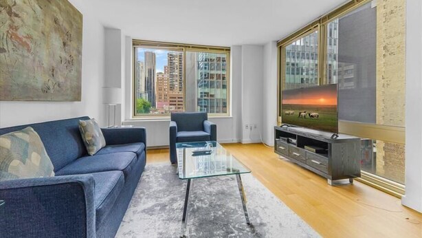Una sala de estar moderna con un sofá, una mesa de centro de vidrio, un televisor sobre un mueble bajo y un ventanal con vista a la ciudad.