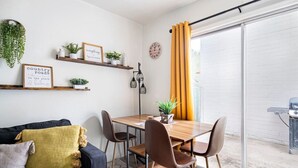 Ein gemütlicher Essbereich mit einem Holztisch, braunen Stühlen und einem schwarzen Sofa mit gelbem Kissen. An der Wand befinden sich Regale mit Pflanzen und gerahmte Schilder.