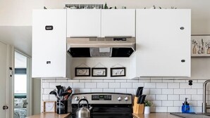 Eine moderne Küche mit weißen Schränken, einem Edelstahlbackofen und einer schwarzen Kochplatte. Über der Kochplatte befinden sich gerahmte Bilder, und links ist ein kleines Fenster.