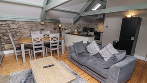 Una sala de estar acogedora con un sofá gris, una mesa de centro de madera y sillas blancas. Se aprecia una cocina al fondo, con una pared de piedra y un refrigerador negro.