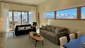 Ein großzügiges Wohnzimmer mit Sofa, Couchtisch und Essbereich. Große Fenster bieten Blick auf ein verschneites Gebirge.