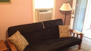A black sofa with two patterned cushions, a wooden side table, a floor lamp, and a door with a warning sign. - Cabin in Bankhead National Forest (Moulton)