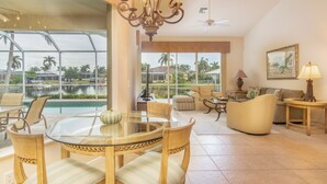 A spacious living area with a glass table, comfortable seating, a ceiling fan, and large windows offering a view of a pool and palm trees.