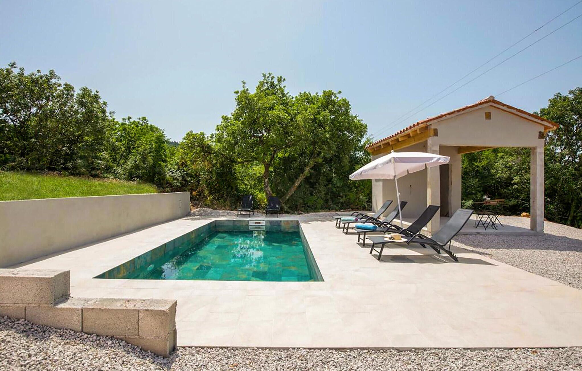 Una piscina rectangular con agua azul cristalina, rodeada por una solana de hormigón. Hay sillas de descanso y un pequeño pabellón con una sombrilla blanca.