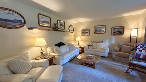 Ein Wohnzimmer mit zwei weißen Sofas, einem Glastisch und einem Holzstuhl. Auf Beistelltischen stehen zwei Lampen, und der Boden ist mit Teppich ausgelegt.