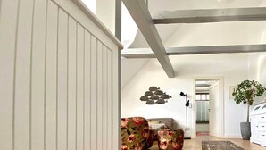 A spacious room with wooden flooring, a white wall, and a ceiling with exposed beams. There is a sofa with floral upholstery and a potted plant.