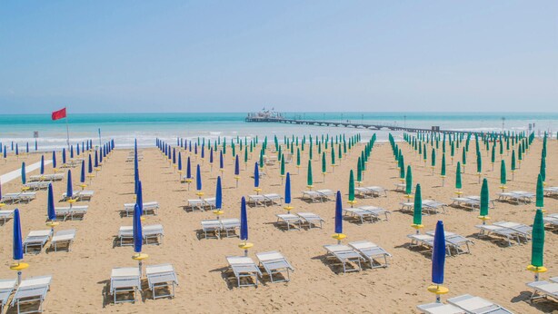 Une plage bordée de rangées de chaises longues et de parasols, une jetée en arrière-plan et un ciel dégagé.