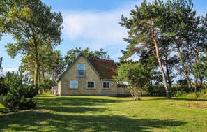Une maison à deux étages, avec une façade en briques et un toit recouvert de tuiles rouges, entourée d’une pelouse bien entretenue et de grands arbres.