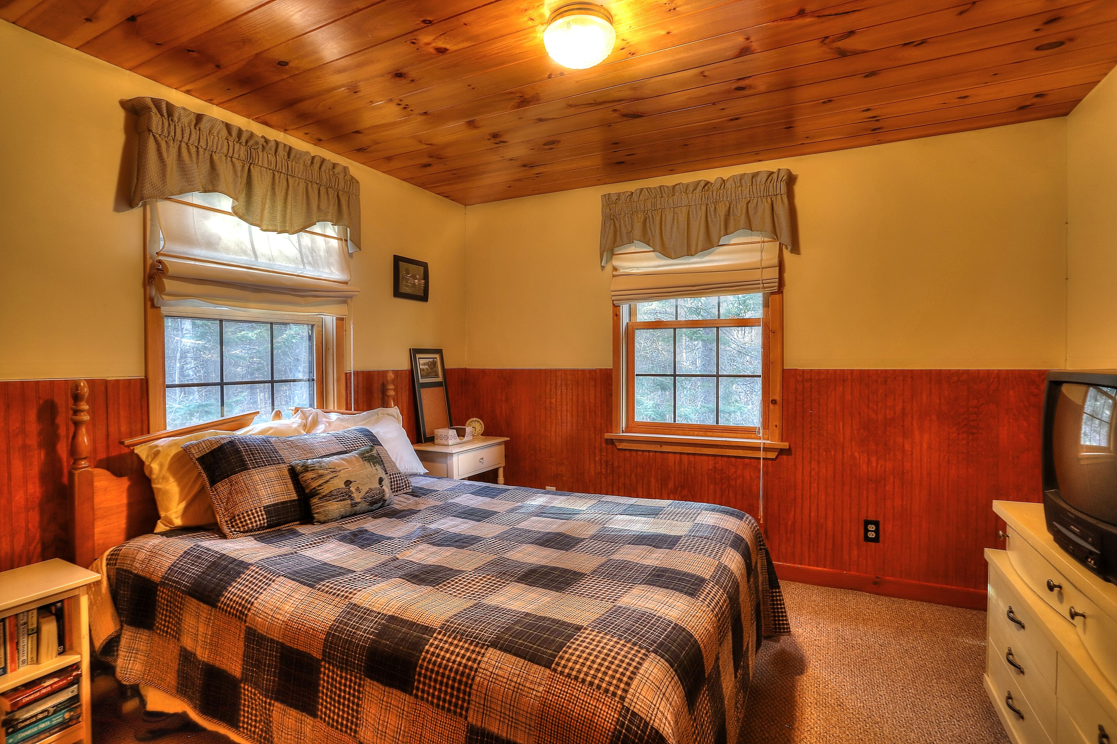 A bedroom with a wooden ceiling, a bed with a checkered quilt, two windows with valances, a nightstand with a lamp, and a dresser with a television.