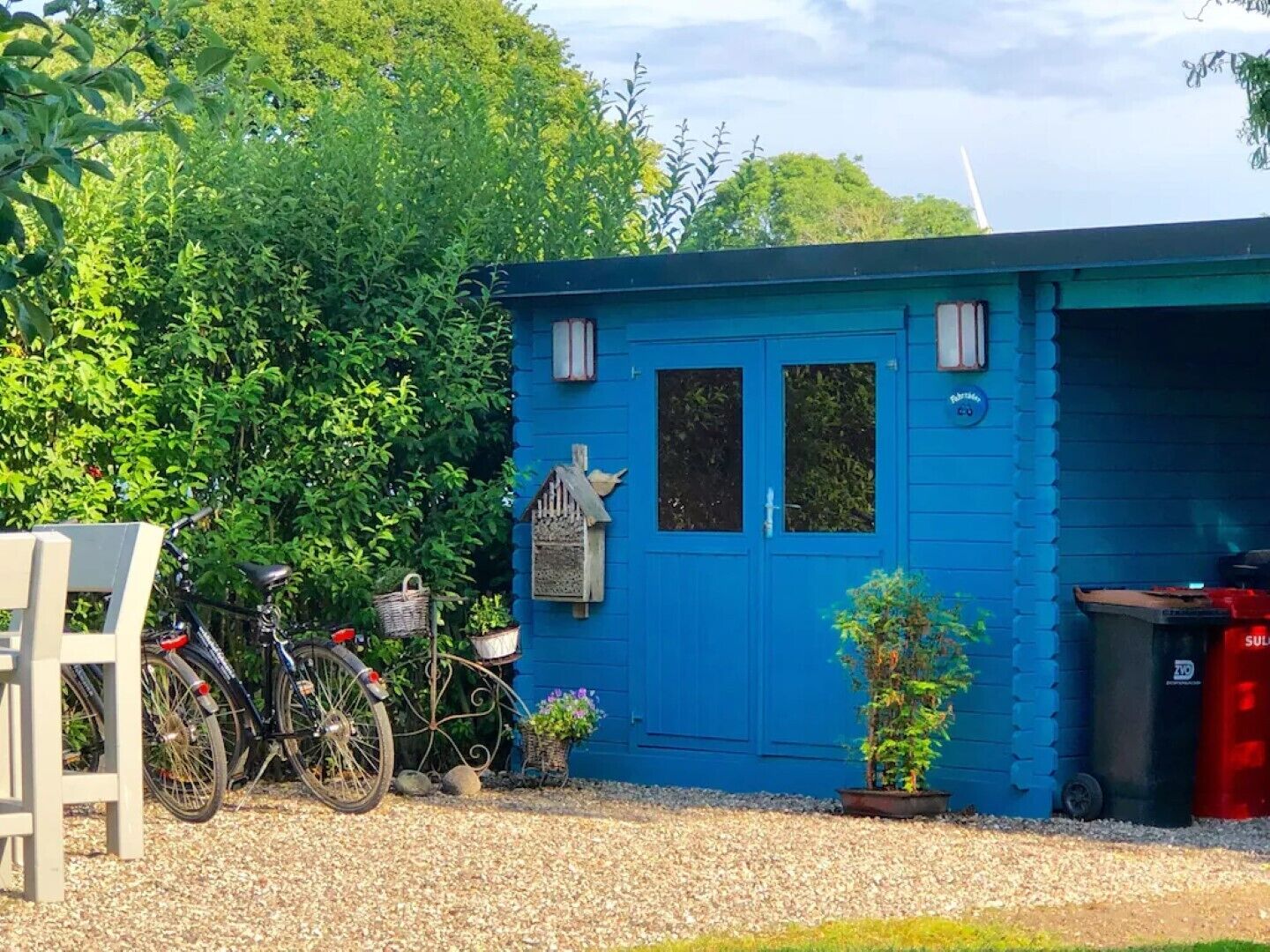 A small blue building with a door and windows, a bicycle, a birdhouse, and a trash bin.