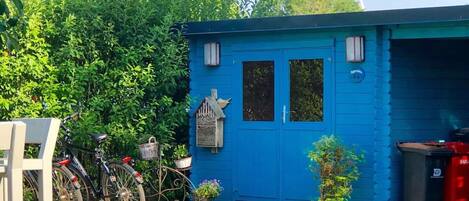 A small blue building with a door and windows, a bicycle, a birdhouse, and a trash bin.
