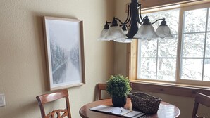 A dining area with a round wooden table, four chairs, a chandelier, a framed picture on the wall, and a window with a view of trees. - Coveted location perfectly close to town, golf, lake & park with surplus parking (McCall)