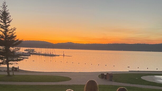 Three people sitting on a grassy area overlooking a body of water with boats and a dock in the background. - Coveted location perfectly close to town, golf, lake & park with surplus parking (McCall)