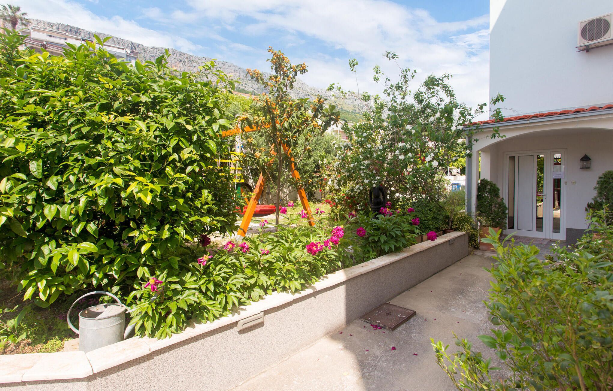 Ein Garten mit verschiedenen Pflanzen und Blumen, einem Metall-Gießkannen und einem weißen Gebäude mit rotem Dach.