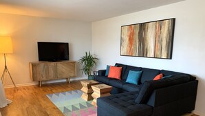 Un salon moderne avec un canapé d’angle, un téléviseur à écran plat, un meuble en bois et un tapis coloré.