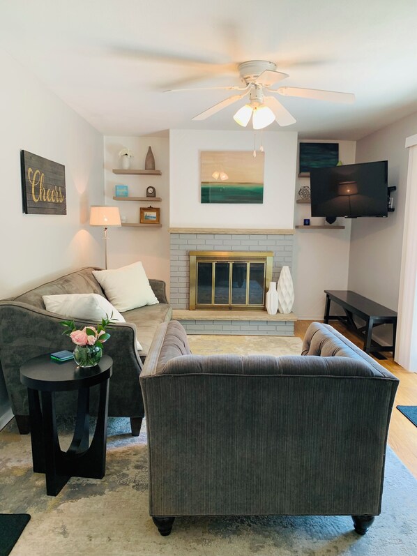 A living room with a fireplace, a ceiling fan, a TV mounted on the wall, and a sofa. - Welcome to Holiday House! Cozy, modern home 7 min. from the beach and wineries. (Stevensville)