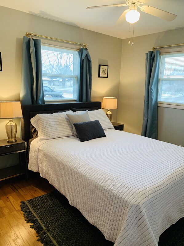 A neatly made bed with a white quilt, two pillows, and a dark headboard. There are two bedside tables with lamps, a dark rug, and a ceiling fan.
