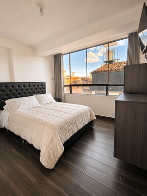 A hotel room with a large bed, a dark wood floor, a view of the cityscape through large windows, and a flat-screen TV mounted on the wall. - MONACO HOUSE (Cusco)