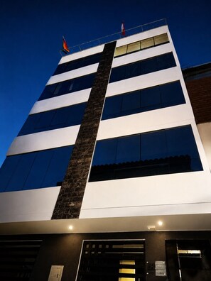 A multi-story building with a prominent vertical black column, large windows, and a flag on top. - MONACO HOUSE (Cusco)