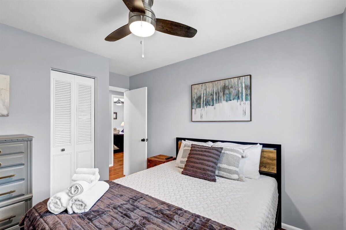 A bedroom with a bed, a ceiling fan, a painting, and a door.