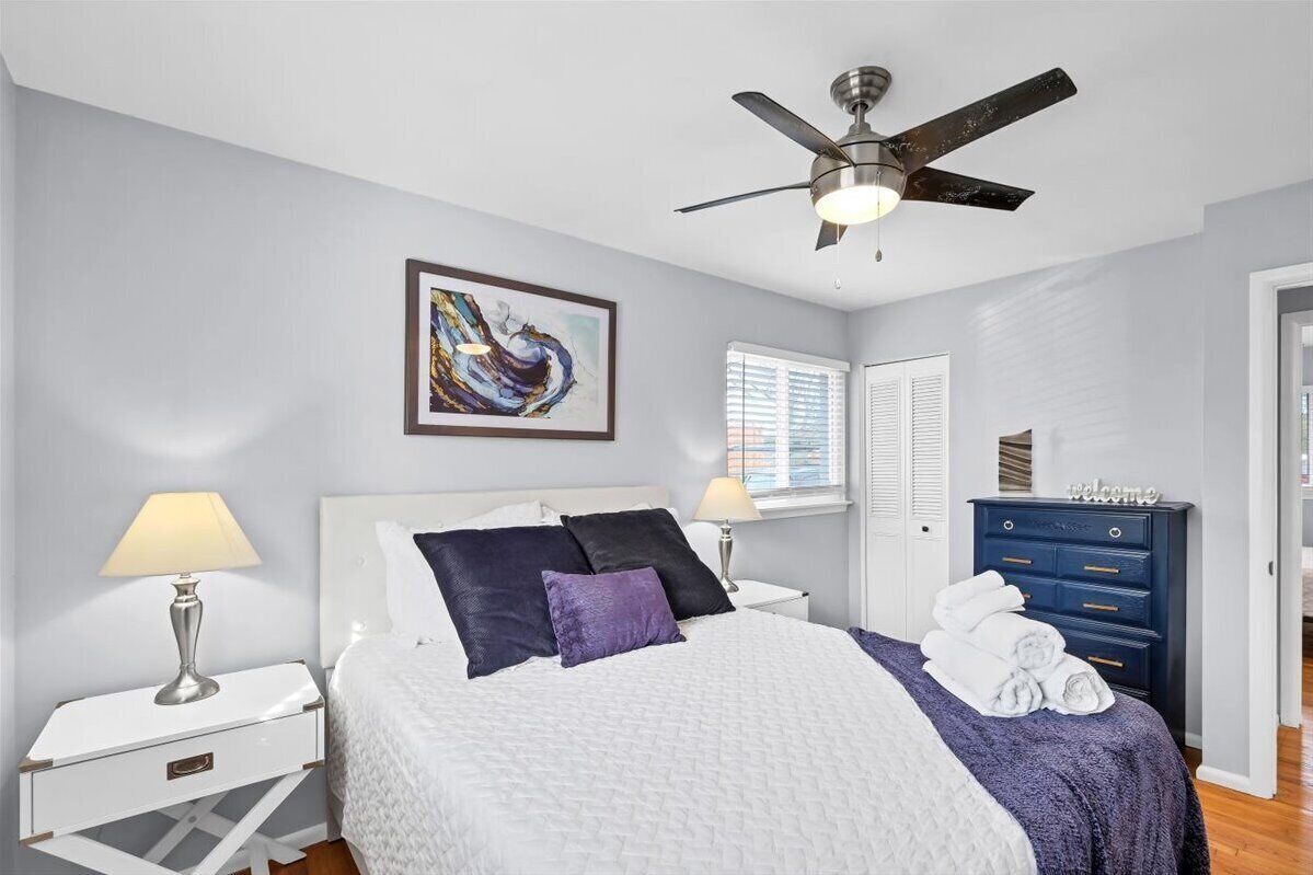 A bedroom with a bed, a nightstand, a dresser, a ceiling fan, a window with blinds, and a framed abstract painting on the wall.