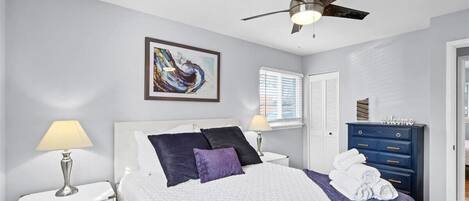 A bedroom with a bed, a nightstand, a dresser, a ceiling fan, a window with blinds, and a framed abstract painting on the wall.