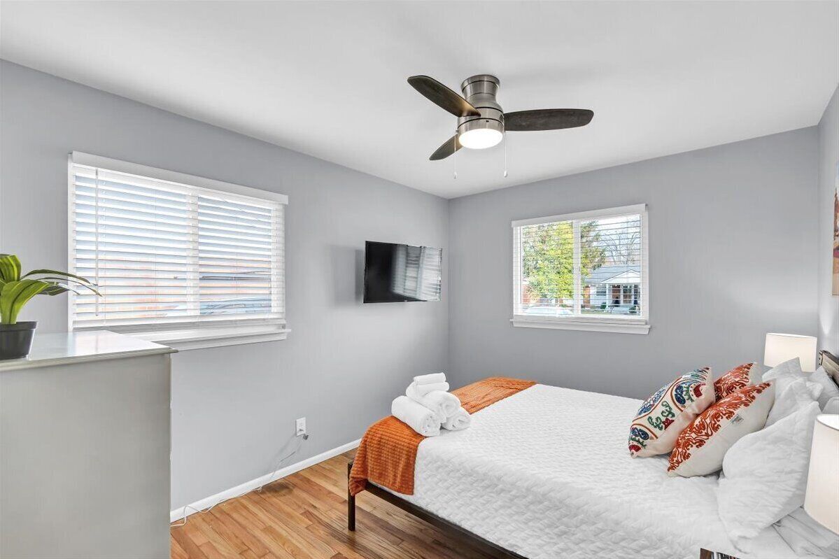 A bedroom with a bed, a ceiling fan, a TV, and windows with blinds.