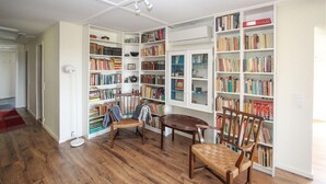 A room with wooden flooring, a bookshelf filled with books, a table with chairs, and a door in the background. - Awesome home in Samsø with sauna (Samsø)