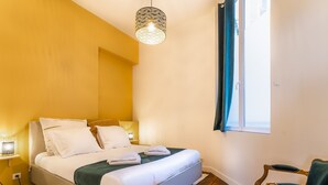Una cama bien tendida con sábanas blancas y un plaid verde, piso de madera, una silla con cojín verde y una lámpara colgante.