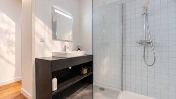 Casa de banho moderna com chuveiro em cabine de vidro, toucador escuro com espelho e lavatório branco.