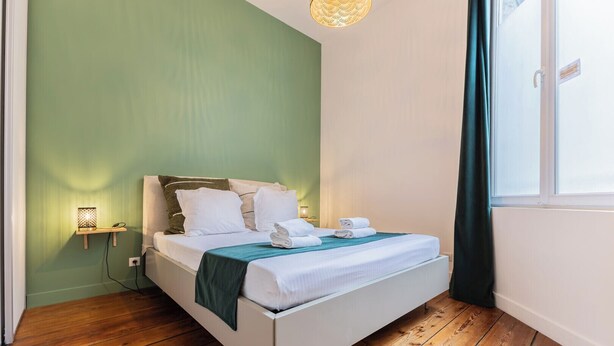 Een net opgemaakt bed met witte lakens en een blauwgroene plaid, een bedlampje en een raam met gordijn.