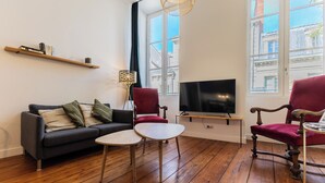 Een woonkamer met een bank, twee fauteuils, een bijzettafeltje, een televisie en een boekenkast.