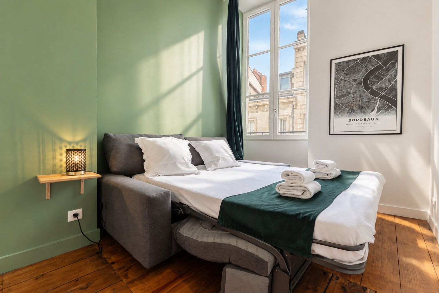 A compact hotel room with a bed, a bedside table with a lamp, a window with a view of buildings, and a framed map on the wall.