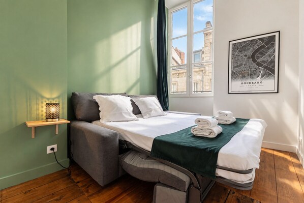 A compact hotel room with a bed, a bedside table with a lamp, a window with a view of buildings, and a framed map on the wall.