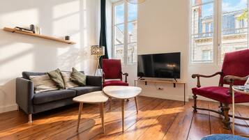 Uma sala de estar com um sofá, duas cadeiras, uma televisão e piso de madeira.