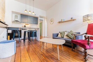 A modern living room with a wooden floor, a sofa, a red chair, a small table, and a kitchen area with a stove and bar stools.