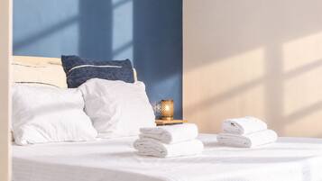 Una cama bien tendida con sábanas blancas, una pared azul y una mesita con una lámpara y toallas dobladas.