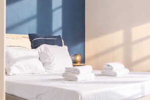 Una cama bien tendida con sábanas blancas, una pared azul y una mesita con una lámpara y toallas dobladas.