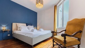 A bedroom with a bed, a chair, a bedside table with a lamp, and a window.