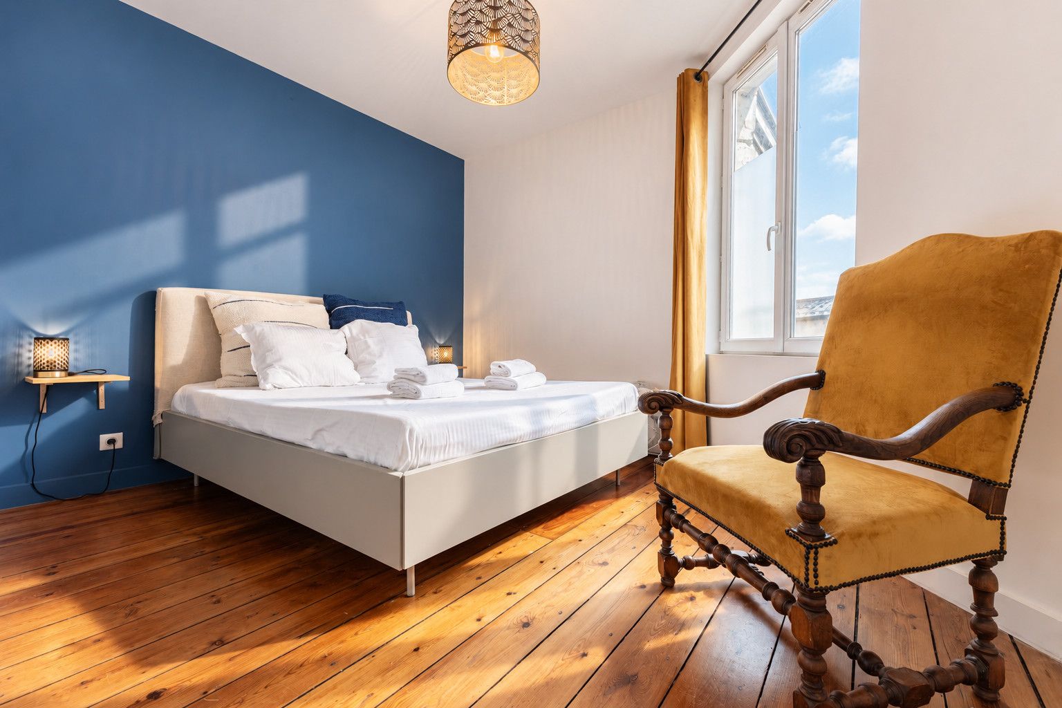 A bedroom with a bed, a wooden floor, a chair, a bedside table with a lamp, and a window with curtains.