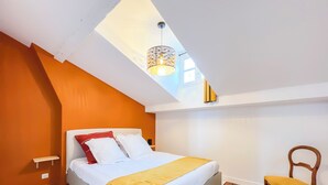 Ein Schlafzimmer mit einem Bett, einem Holzsessel, einem kleinen Nachttisch und einem Oberlicht.