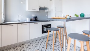 A modern kitchen with white cabinets, a black countertop, and a tiled backsplash. There are two stools and a small table with a basket of apples and a green vase.