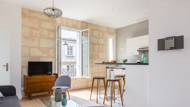 A modern living room with a glass coffee table, grey sofas, a TV on a wooden cabinet, and a kitchen area with stools and a small island.
