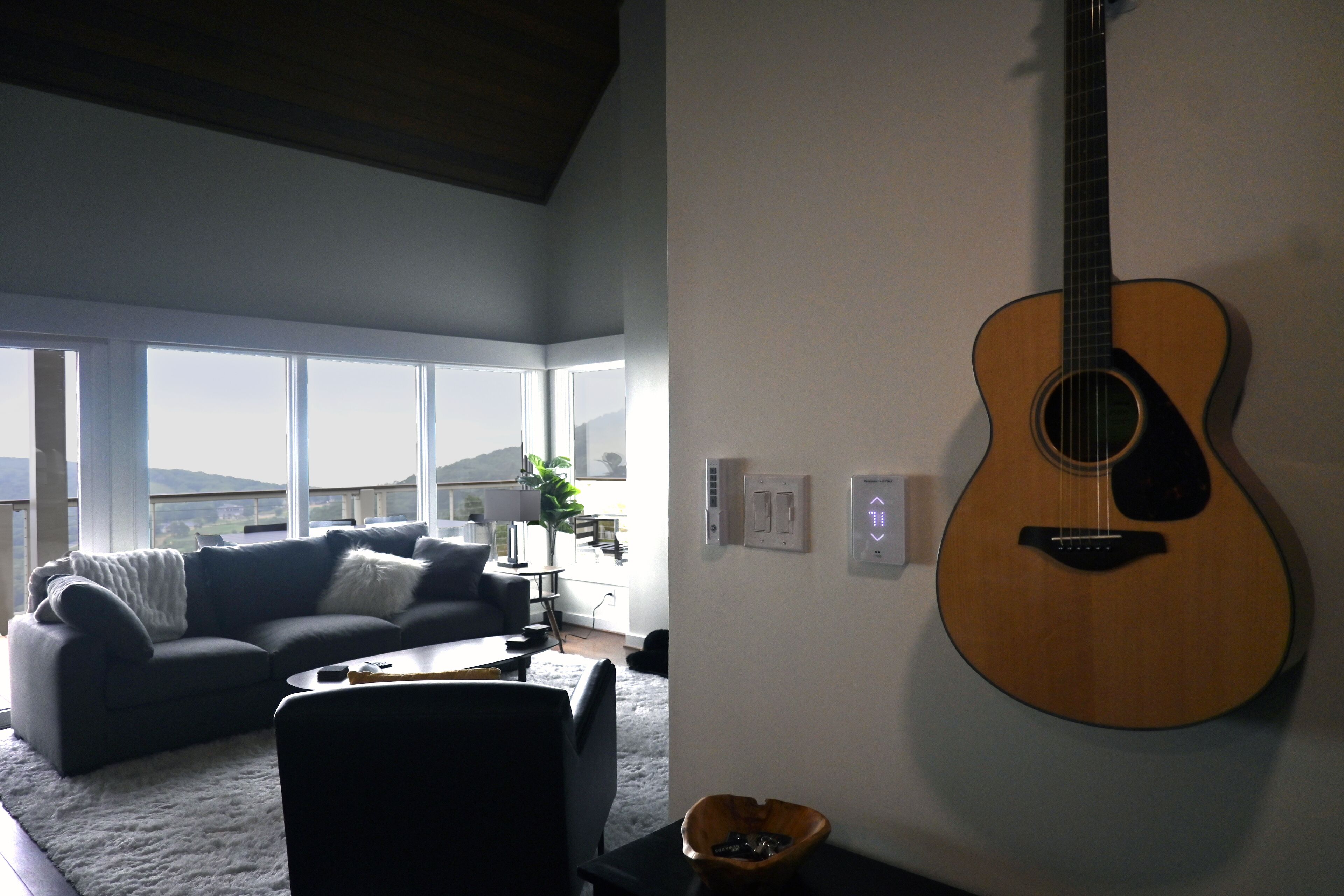 Een woonkamer met een groot raam, een grijze bank, een bijzettafeltje en een gitaar aan de muur.