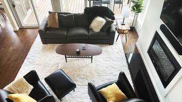 Un salon moderne avec un canapé de couleur foncée, deux fauteuils en cuir noir, un tapis à poils longs blanc et un téléviseur à écran plat fixé au mur.