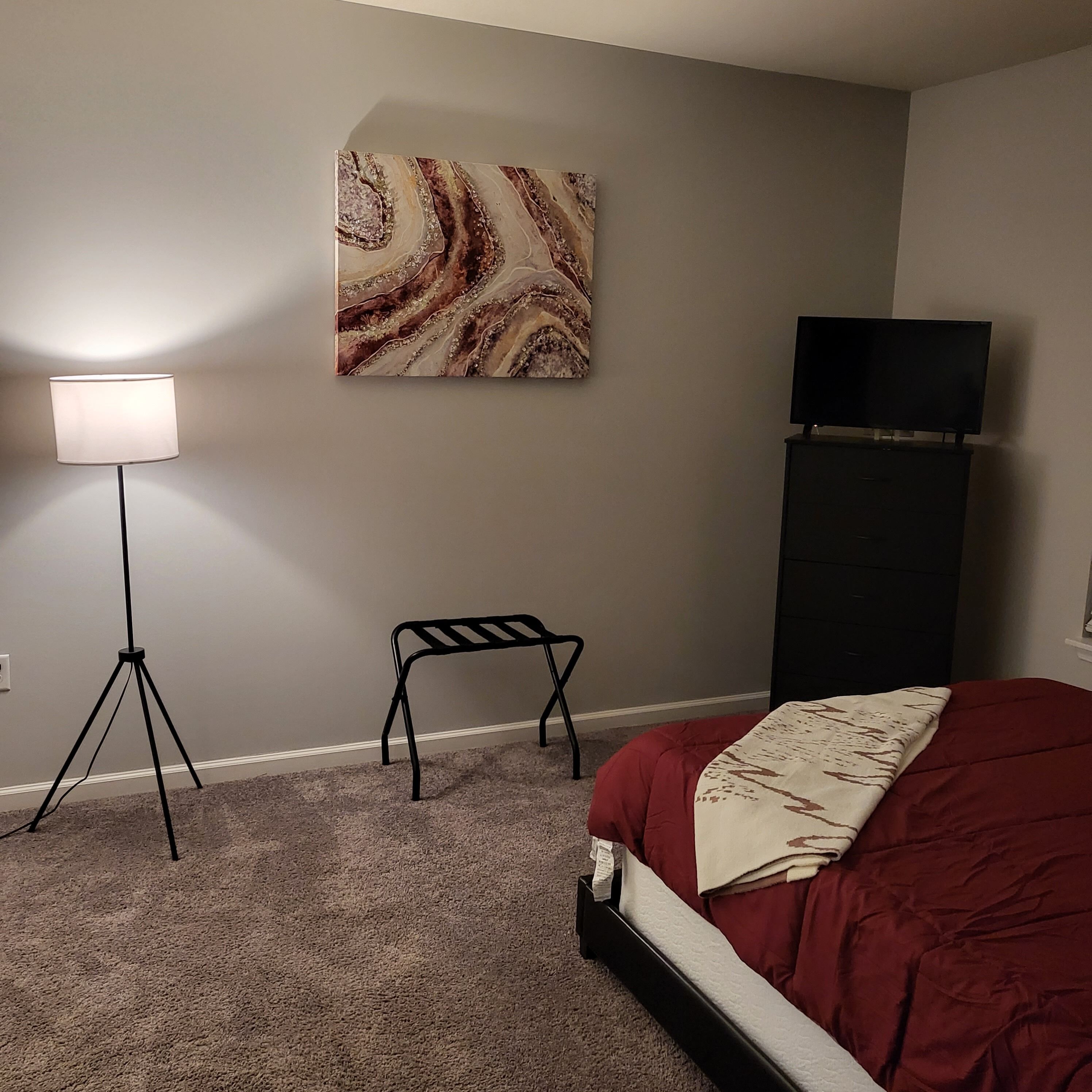A bedroom with a bed, a floor lamp, a black chair, a TV on a stand, and a marble-patterned artwork on the wall.