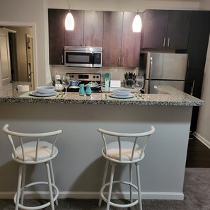A kitchen with dark wooden cabinets, a stainless steel refrigerator, a microwave, and a stove. There are two white bar stools and a granite countertop. - Stylish and Spacious, Close to the Hospital (Fort Wayne)